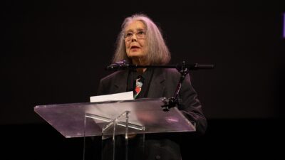 Poet Anne Carson speaking at a lectern at the 2024 National Book Awards finalist reading in New York