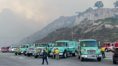 Fire engines and crews staged along a coastal road beneath smoke-covered hills in Southern California